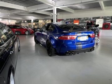 In a garage stands among other vehicles a blue sports car with spoiler and black rims, proudly bearing license plates. Part of the Jaguar Heritage Trust Collection, it stands out against the background decorated with vintage photographs.