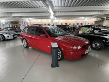 A red station wagon from the Jaguar Heritage Trust collection is displayed with other vehicles in a bright, spacious showroom. It is slightly angled, with informative signage at the front.