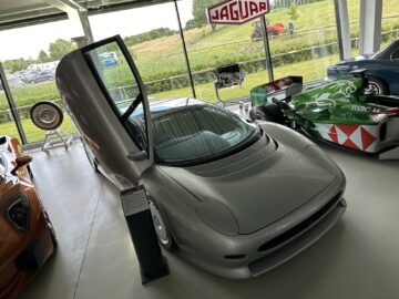 A silver sports car with open scissor doors, displayed alongside other vehicles in a showroom with large windows, is part of the Jaguar Heritage Trust Collection. A green formula racing car is visible in the background, which adds a touch of historic elegance to this car report.