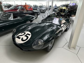 A green vintage race car with the number 25 on the hood is on display in a showroom full of other classic vehicles, part of the illustrious Jaguar Heritage Trust Collection.