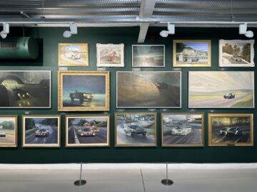 A gallery wall featuring a collection of car-themed framed paintings, all vividly depicting cars and racing scenes. The frames are arranged in three rows against a green background, with highlights from the Jaguar Heritage Trust.