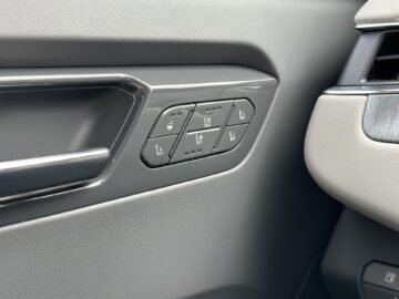 Close-up van het binnendeurpaneel van de Kia EV9 uit 2024, met verschillende stoelverstelknoppen, waaronder verwarmings- en koelfuncties. Het voornamelijk grijze paneel met metallic accenten weerspiegelt het premium gevoel van dit RWD-wonder.