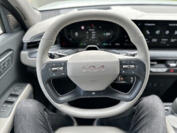 Close-up van een Kia EV9-stuur uit 2024, met verschillende bedieningsknoppen, een digitaal dashboard en een deel van de schoot van de bestuurder in deze geavanceerde RWD-SUV.