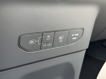 Een close-up van het dashboardpaneel van de Kia EV9 uit 2024, met USB-poorten, een 12V-stopcontact en een drukknop.