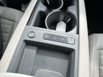 Close-up van de middenconsole van de Kia EV9 uit 2024 met een bekerhouder, een "Auto Hold"-knop, een klimaatcontroleknop, een parkeerhulpknop en een gestructureerd compartiment voor kleine spullen in de RWD-indeling.