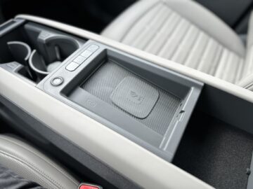 Close-up van de middenconsole van een Kia EV9 uit 2024 met een oplaadpad voor draadloze telefoons, bekerhouders en bedieningsknoppen. De omgeving omvat een lichtgekleurde stoel en een armleuning, die het strakke interieurdesign van het RWD-model illustreren.