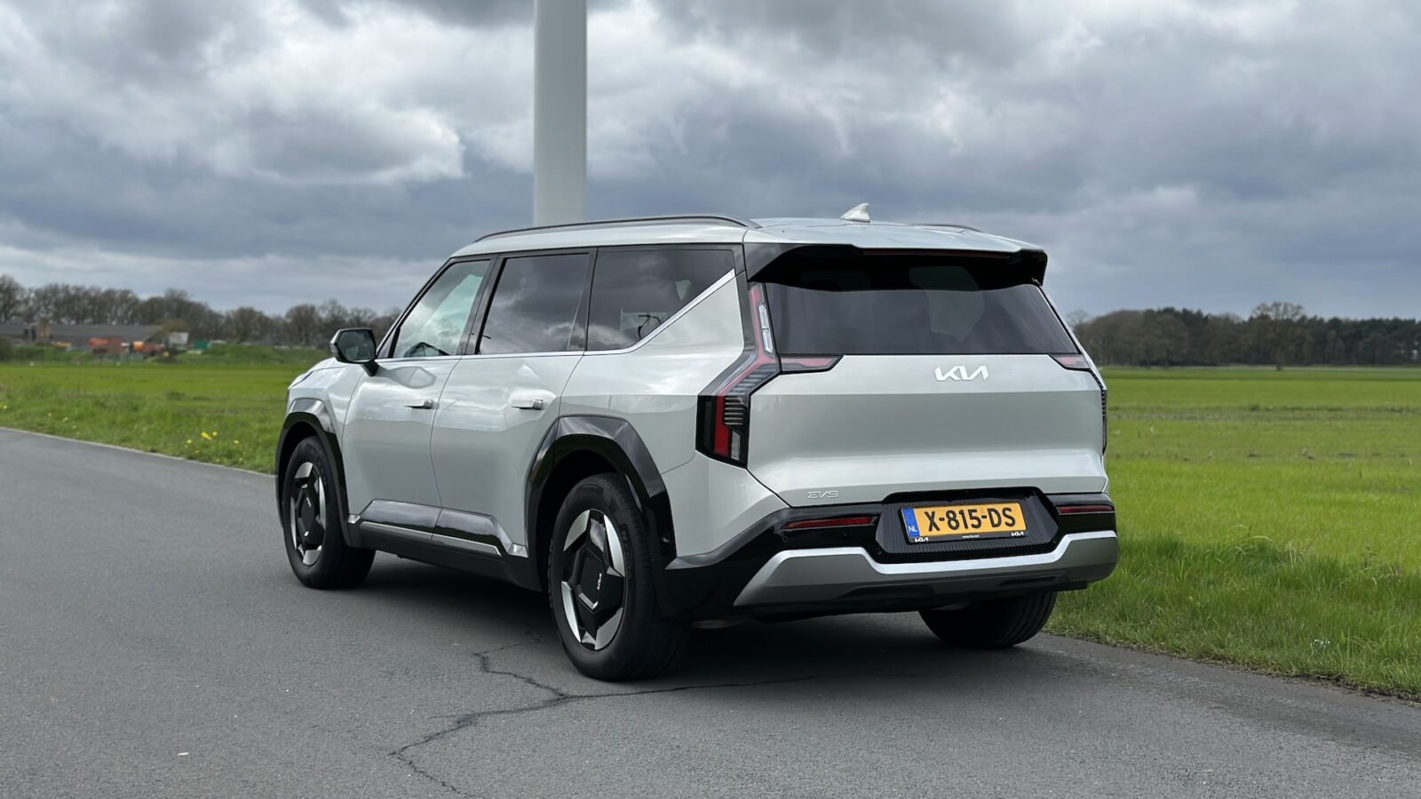 Een strakke zilverkleurige Kia EV9 uit 2024 met Nederlands kenteken staat geparkeerd aan de kant van een landelijke weg, omlijst door een groot veld en een bewolkte lucht op de achtergrond.