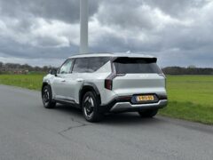 Een strakke zilverkleurige Kia EV9 uit 2024 met Nederlands kenteken staat geparkeerd aan de kant van een landelijke weg, omlijst door een groot veld en een bewolkte lucht op de achtergrond.