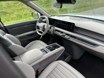 Een blik op het Kia EV9-interieur uit 2024 met een wit stuur, een digitaal dashboard, lichtgrijze stoelen en een middenconsole met bekerhouders en bedieningsknoppen. De RWD-auto staat geparkeerd in de buurt van een grasveld.