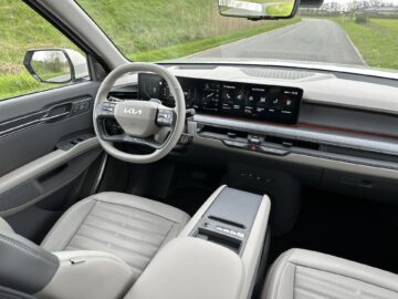 Binnenaanzicht van de moderne cockpit van de Kia EV9 uit 2024, met een stuur, een digitaal dashboard en een centraal touchscreen-display, met een weg zichtbaar door de voorruit. Dit RWD-voertuig combineert geavanceerde technologie met een strak ontwerp voor een ongeëvenaarde rijervaring.