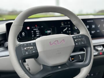 Close-up van een Kia EV9-autostuur uit 2024, met verschillende bedieningsknoppen en een deel van het digitale dashboarddisplay op de achtergrond.