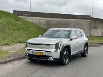 Een zilverkleurige Kia EV9 SUV uit 2024 staat geparkeerd op een weg naast een grasveld en een betonnen muur onder een bewolkte hemel. Op het kenteken aan de voorkant staat "X-815-DS.