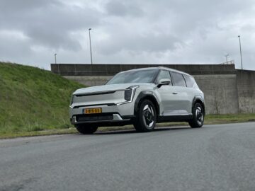 Een witte Kia EV9 uit 2024 met zwarte details staat geparkeerd op een verharde weg nabij een met gras begroeide helling en een betonnen muur onder een bewolkte hemel.