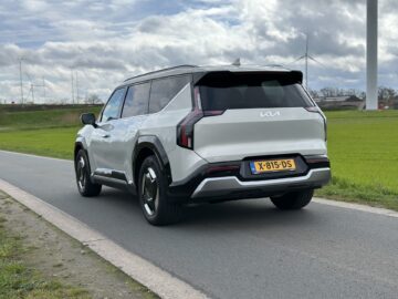 Een zilveren Kia EV9 SUV uit 2024 staat geparkeerd op een verharde weg naast een grasveld onder een bewolkte hemel.