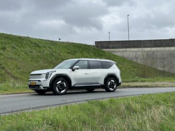 Een witte Kia EV9 SUV uit 2024 staat op een bewolkte dag geparkeerd op een weg naast een met gras begroeide helling, met een betonnen structuur op de achtergrond. Dit RWD-model belichaamt zowel modern design als geavanceerde technologie.