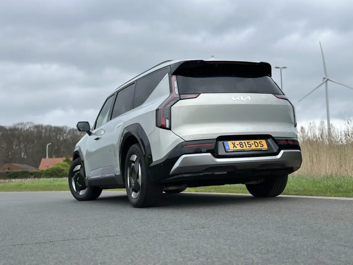 Een witte Kia EV9 SUV uit 2024 met een zichtbaar kenteken "X-815-DS" staat geparkeerd op een weg naast een veld met windturbines op de achtergrond onder een bewolkte hemel.