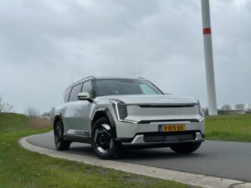 Op een verharde weg staat een zilveren Kia EV9 uit 2024 met Nederlands kenteken geparkeerd. De achtergrond omvat groen en een windturbine, onder een bewolkte hemel.