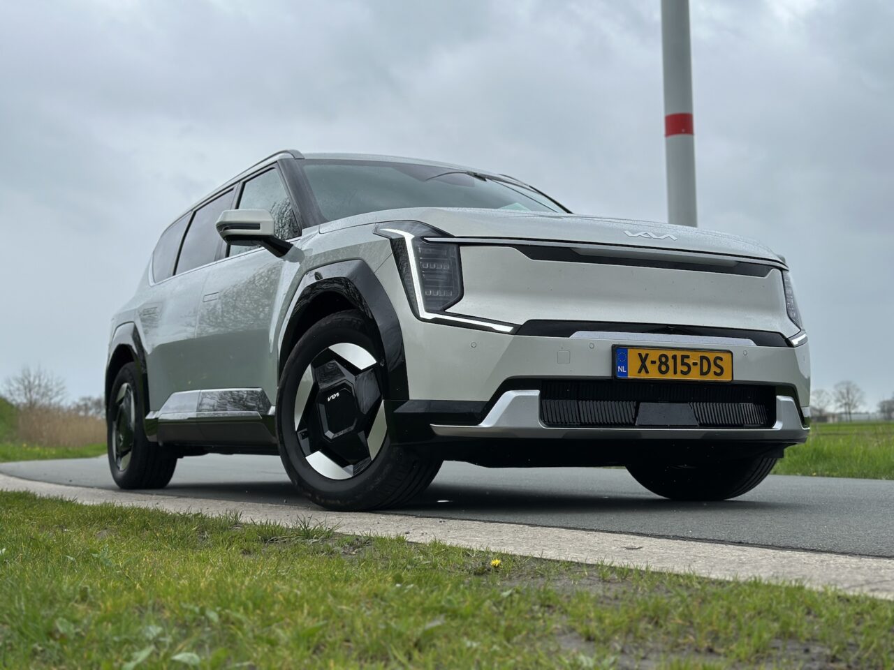 Een zilverkleurige Kia EV9-SUV uit 2024 staat op een bewolkte dag langs de kant van de weg geparkeerd, met op het Nederlandse kenteken "X-815-DS".