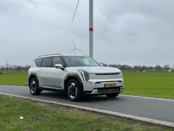 Een strak witte Kia EV9 uit 2024 staat geparkeerd aan de kant van een landelijke weg, met sierlijk draaiende windturbines op de achtergrond.