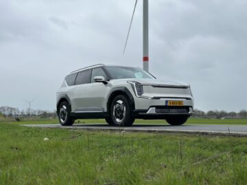 Een zilverkleurige Kia EV9 SUV uit 2024, met achterwielaandrijving, staat geparkeerd op een weg met een windturbine op de achtergrond. Op het kenteken staat "X-815-DS." De lucht is bewolkt en de omgeving is grasachtig.