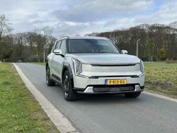 Aan de rechterkant van de weg staat een zilveren Kia EV9 SUV met een Europees kenteken, "X-815-DS", geparkeerd. Bomen en een bewolkte lucht vormen een serene achtergrond voor dit RWD-model uit 2024.