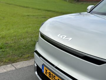 Close-up van de voorkant van een witte Kia EV9 RWD uit 2024 met een zichtbaar logo op de motorkap en gedeeltelijk kenteken "X-815-". De auto staat geparkeerd aan de kant van een weg met een grasveld op de achtergrond.