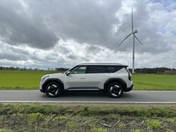 Een Kia EV9 uit 2024, een indrukwekkende witte SUV met achterwielaandrijving, staat geparkeerd op een landweg met een windturbine en groene velden op de achtergrond onder een bewolkte hemel.