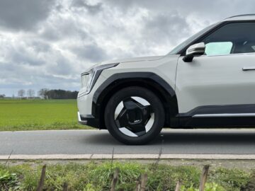 Close-up van de voorkant van een Kia EV9 uit 2024, geparkeerd op een weg, met een grasveld en een bewolkte lucht op de achtergrond. De auto beschikt over strakke, moderne designelementen, zwarte wieldoppen en RWD-dynamiek.