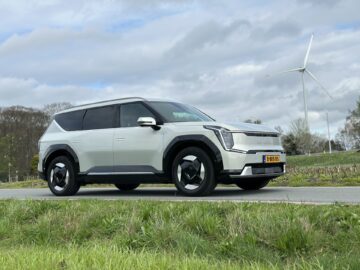 Een witte Kia EV9 uit 2024 staat geparkeerd op een weg in de buurt van groen gras en een windturbine. Het RWD-voertuig heeft een Europees kenteken en de lucht erboven is gedeeltelijk bewolkt.