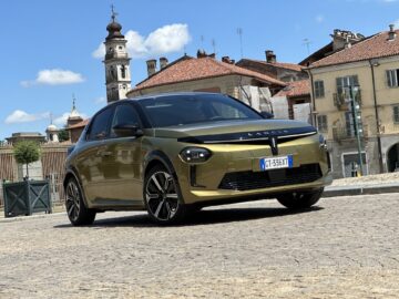 Een goudkleurige Lancia Ypsilon 2024 staat geparkeerd in een geplaveide straat met historische gebouwen en een kerktoren op de achtergrond onder een blauwe hemel, wat zijn tijdloze elegantie en premium belofte laat zien.
