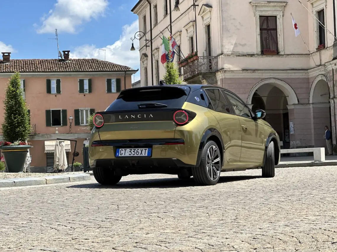 Un Lancia Ypsilon verde, que encarna su promesa premium 2024, está aparcado en un día soleado en una plaza adoquinada con edificios de estilo europeo al fondo.