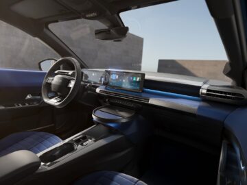 Interieur van de Lancia Ypsilon 2024 met een digitaal dashboarddisplay, een stuurwiel en gewatteerde stoelen met blauwe accenten. Het ontwerp is strak en minimaal, met de nadruk op technologie en comfort, en doet zijn premiumbelofte echt eer aan.