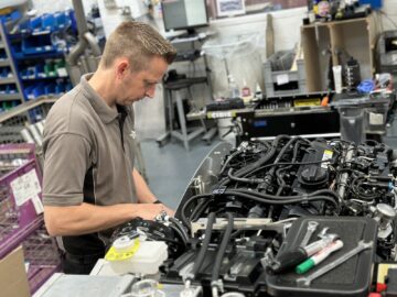 Een man werkt aan een complexe motorassemblage in een georganiseerde werkplaats vol met verschillende gereedschappen en apparatuur, die doet denken aan het nauwgezette vakmanschap bij de Morgan Motor Company.