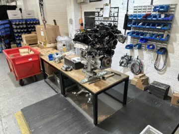 Een nette werkplaats met een werkbank met een motor, gereedschap en onderdelen, geordend in dozen en planken, wat doet denken aan de nauwgezette organisatie bij de Morgan Motor Company.