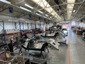 Een werkplaats waar verschillende autochassis door arbeiders worden gemonteerd onder een hoog, helder verlicht plafond. In stellingen tegen de wanden zijn diverse gereedschappen en onderdelen opgeslagen. Een typische dag bij Morgan Motor Company, waar bezoekers kunnen genieten van een gedetailleerde rondleiding door het montageproces.