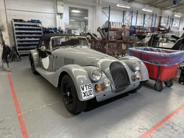 Een klassieke grijze sportwagen met het kenteken "VT19 XJD" staat geparkeerd in een werkplaats, omringd door apparatuur en gereedschap, die doet denken aan een scène uit een Rondleiding Morgan Motor Company-tour of elke plaats waar ze hartstochtelijk auto's maken.