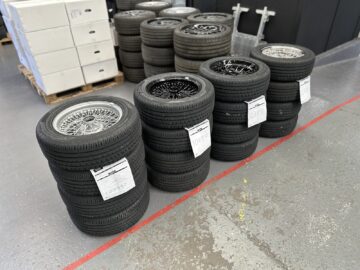 Verschillende stapels banden met aangehechte labels zijn gerangschikt in een garage of werkplaats, wat het nauwgezette vakmanschap oproept dat te zien is tijdens een rondleiding door Morgan Motor Company. Op de achtergrond zijn extra banden en witte dozen te zien, die verwijzen naar de processen die betrokken zijn bij het maken van auto's.