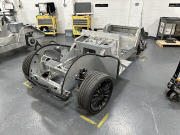 Een metalen voertuigchassis met zwarte wielen staat in een industriële werkplaats, omgeven door geel gereedschap op planken en werkstations tegen witte bakstenen muren. Het lijkt op het vakmanschap dat je ziet bij de Morgan Motor Company.