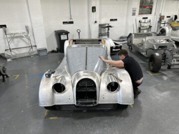 Een persoon werkt aan de voorkant van een gedeeltelijk geassembleerde oldtimer in een werkplaats met op de achtergrond gevulde werkstations die met gereedschap gevuld zijn, wat de klassieke charme van Morgan Motor Company oproept. Als je van rondleidingen houdt, zou dit tafereel de droom van elke autoliefhebber zijn.