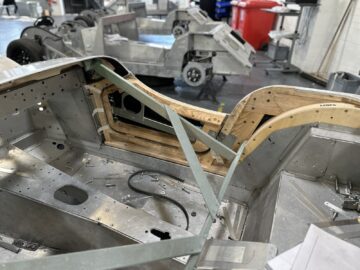 Een close-up van een onafgewerkt autoframe in een werkplaats, met metalen en houten onderdelen. Op de achtergrond zijn andere autoframes en gereedschappen zichtbaar. Deze scène geeft de essentie weer van de Rondleiding Morgan Motor Company, waar toegewijde ambachtslieden zelden auto's met de hand vervaardigen.