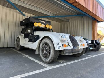 Een cabriolet in vintage-stijl van Morgan Motor Company, voorzien van offroad-banden en een daktent, staat geparkeerd in een overdekte ruimte met kentekenplaat VK22 NFR. Deze unieke auto wacht op zijn volgende avontuur of misschien zelfs op een rondleiding die zijn klassieke charme laat zien.