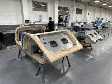 Workshopscène met carrosserieframes, één met een houten buitenstructuur en de andere met een metalen frame, geplaatst op standaards. Op de achtergrond is een persoon in een donkere outfit te zien, wat doet denken aan het vakmanschap bij Morgan Motor Company.