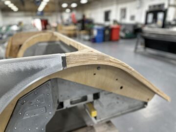 Een close-up van een onafgewerkte houten en metalen structuur met een gebogen verbinding, gelegen in een werkplaats met wazige gereedschappen en apparatuur op de achtergrond, die doet denken aan het vakmanschap bij Morgan Motor Company.