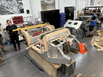 Mensen namen deel aan een workshop over de constructie van autochassis met verschillende gereedschappen en materialen verspreid, wat doet denken aan een tafereel dat je tegenkomt tijdens een rondleiding bij de historische Morgan Motor Company.