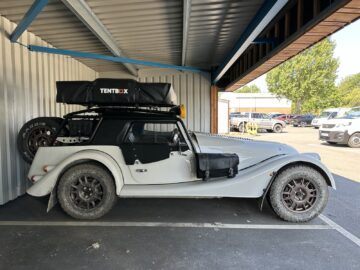 Een witte, vintage-stijl Morgan Motor Company-auto uitgerust met offroad-banden, een TENTBOX-daktent en een reservewiel staat geparkeerd in een overdekte ruimte naast een zilveren golfplaten wand.