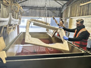 Twee arbeiders in veiligheidsvesten en handschoenen laten een houten frame in een grote container gevuld met een rode vloeistof zakken in een industriële faciliteit, wat doet denken aan het nauwgezette vakmanschap dat te zien was tijdens een Rondleiding bij de Morgan Motor Company.