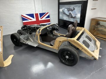 Voertuigframe met zichtbaar chassis en houten panelen, met twee stoelen, stuurwiel en Union Jack-vlag op de achtergrond: een ware knipoog naar het vakmanschap van de klassieke auto's van Morgan Motor Company.