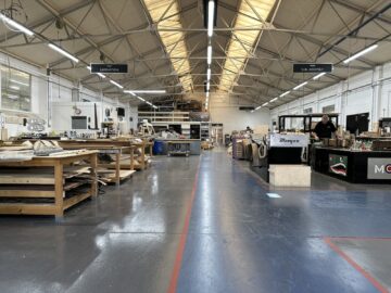 Een ruime fabrieksvloer met hoge plafonds en diverse werkplekken. Eén persoon werkt aan een zwarte machine met het opschrift 'Morgan', die doet denken aan het vakmanschap bij Morgan Motor Company. Aan het plafond hangen bordjes met de tekst ‘Lamination’ en ‘Sub-Assembly’, die bezoekers begeleiden tijdens hun rondleiding.