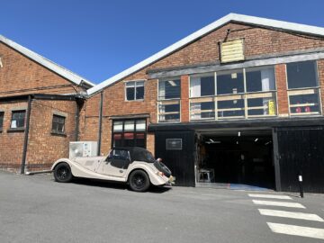 Een vintage cabriolet van Morgan Motor Company staat geparkeerd buiten een bakstenen industrieel gebouw met een open garagedeur. De lucht is helder en zonnig, waardoor het perfect weer is voor een autorondleiding.