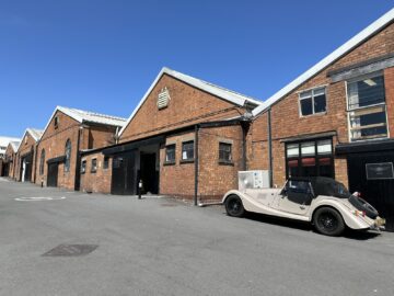 Op een heldere, zonnige dag staat een klassieke witte auto geparkeerd voor een rij bruine bakstenen industriële gebouwen, die doen denken aan een Rondleiding Morgan Motor Company waar zelden auto's met precisie worden vervaardigd.